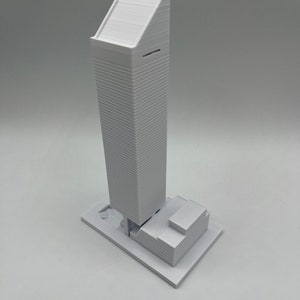 May include: A white 3D printed model of a skyscraper with a flat roof and a smaller building at the base.