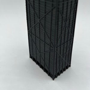 May include: A black, 3D-printed architectural model of a skyscraper. The model features intricate details of the building's facade, including vertical and diagonal support beams. The model is set against a white background.