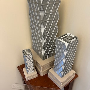 May include: Three grey 3D printed models of skyscrapers with a geometric design. The models are arranged in a row on a wooden table with a ruler in the foreground.