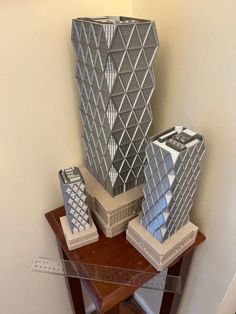 May include: Three grey 3D printed models of skyscrapers with a geometric design. The models are arranged in a row on a wooden table with a ruler in the foreground.