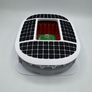 May include: A miniature stadium model with a white exterior, a black grid roof, and a green field. The stadium has a red interior with vertical yellow lines. The model is on a white base.