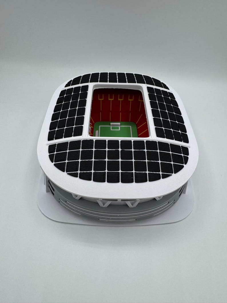 May include: A miniature stadium model with a white exterior, a black grid roof, and a green field. The stadium has a red interior with vertical yellow lines. The model is on a white base.