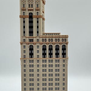 May include: A detailed model of a tall building with a beige facade, featuring numerous windows and architectural details. The building has a tower section and is accented with brown trim. The model is a miniature representation of a skyscraper.