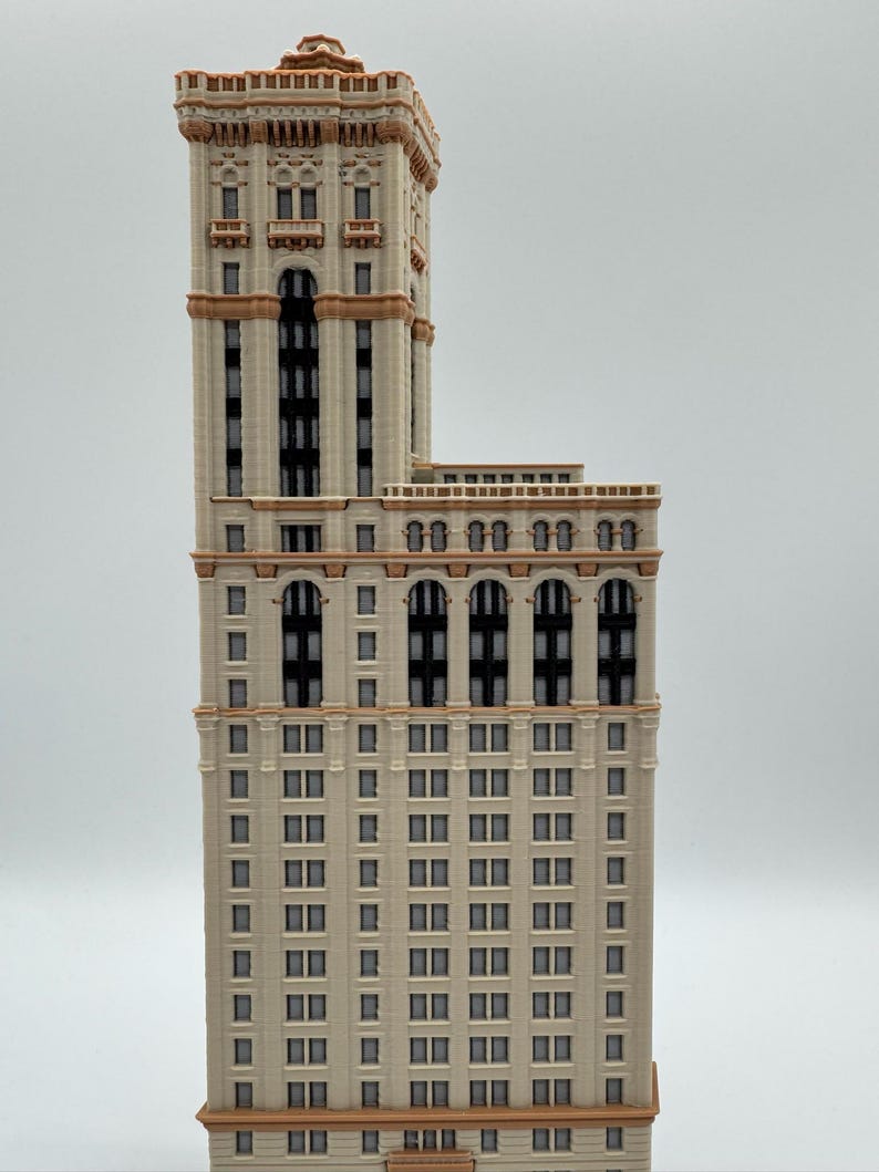 May include: A detailed model of a tall building with a beige facade, featuring numerous windows and architectural details. The building has a tower section and is accented with brown trim. The model is a miniature representation of a skyscraper.