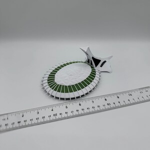 Puede incluir: Maqueta de papel blanca y verde de un estadio, con una regla al lado. El modelo del estadio tiene forma circular con detalles blancos y verdes. La regla est&aacute; marcada con cent&iacute;metros.
