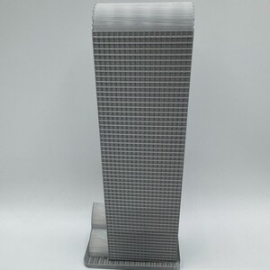 May include: A gray, miniature replica of a skyscraper. The building has a rectangular shape with a textured facade of numerous small, square windows. The top of the building has a textured, flat roof. The base of the building is also gray and rectangular.