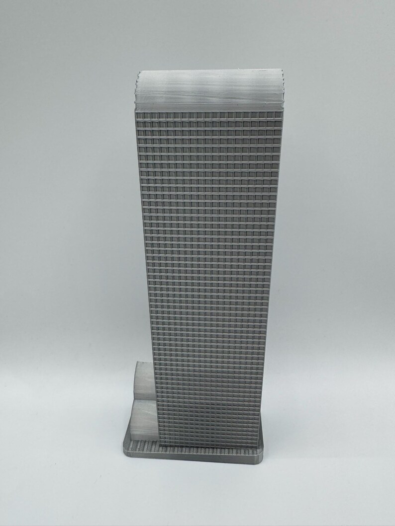 May include: A gray, miniature replica of a skyscraper. The building has a rectangular shape with a textured facade of numerous small, square windows. The top of the building has a textured, flat roof. The base of the building is also gray and rectangular.