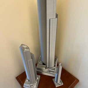 May include: Three gray 3D printed models of skyscrapers, the tallest is about 12 inches tall, the middle is about 8 inches tall, and the smallest is about 5 inches tall. The models are on a wooden table with a ruler.