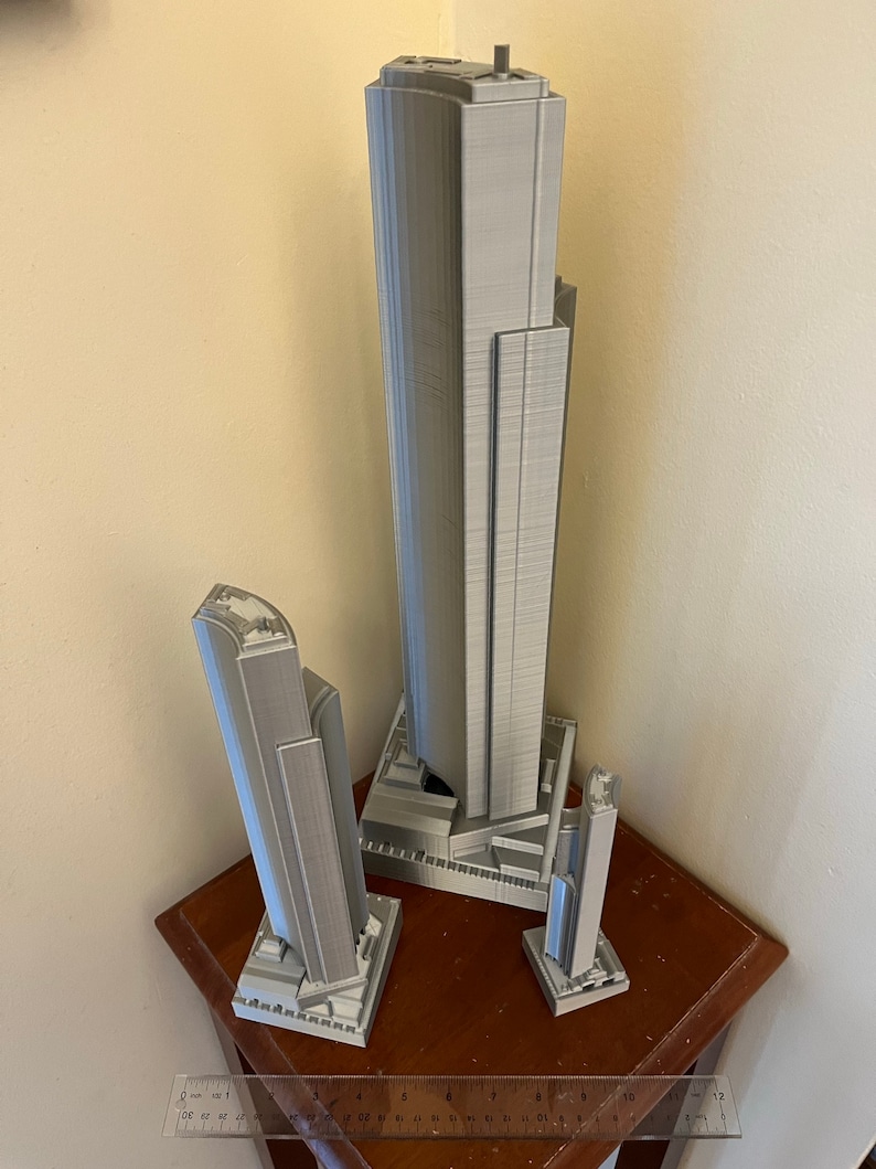 May include: Three gray 3D printed models of skyscrapers, the tallest is about 12 inches tall, the middle is about 8 inches tall, and the smallest is about 5 inches tall. The models are on a wooden table with a ruler.