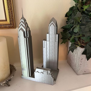 May include: A silver 3D printed model of two skyscrapers. The model is on a shelf with a candle and a potted plant.
