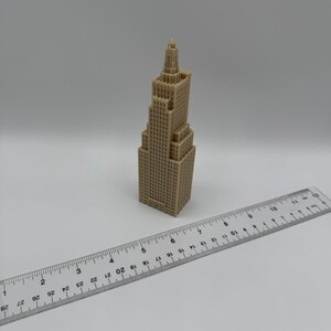 May include: A tan-colored miniature skyscraper model with a detailed facade. A clear ruler is placed next to the building, showing measurements in inches. The model has a tiered design and a spire at the top.