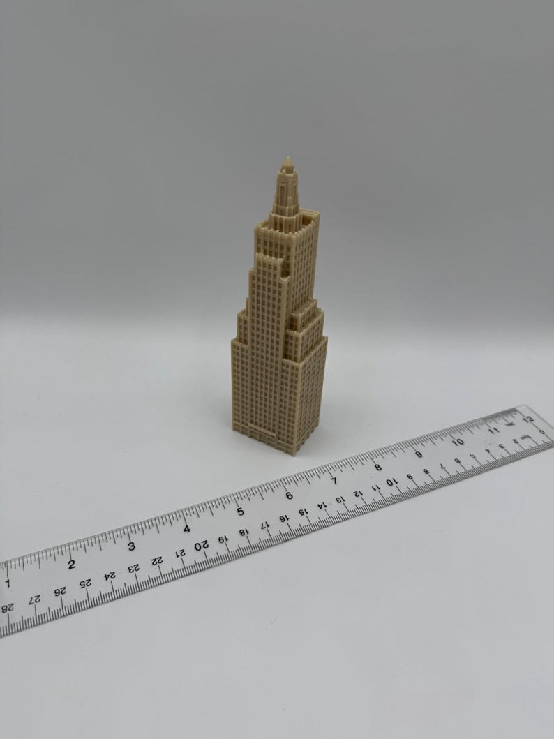 May include: A tan-colored miniature skyscraper model with a detailed facade. A clear ruler is placed next to the building, showing measurements in inches. The model has a tiered design and a spire at the top.