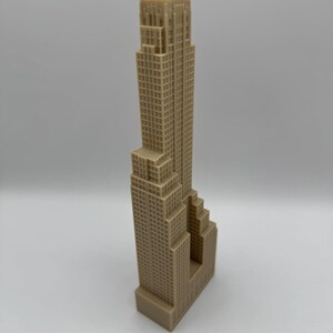 May include: A tan-colored miniature skyscraper model with a detailed facade. The building has a rectangular base and a tall, multi-tiered structure with numerous windows. The model is a representation of a famous building.