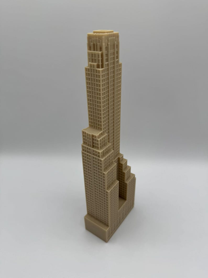 May include: A tan-colored miniature skyscraper model with a detailed facade. The building has a rectangular base and a tall, multi-tiered structure with numerous windows. The model is a representation of a famous building.
