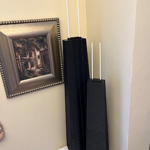 May include: Two black architectural models of the Willis Tower, formerly the Sears Tower, with white antenna-like structures extending from the top. The models are displayed on a white surface against a beige wall, with a framed picture in the background.
