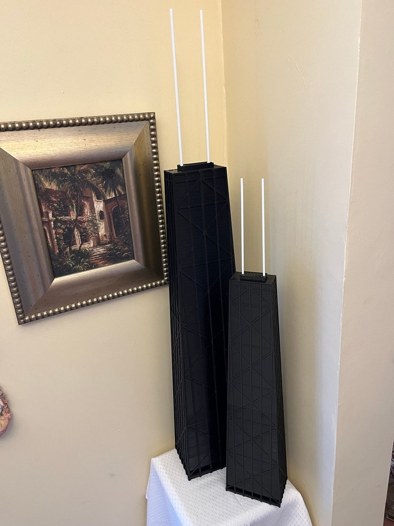 May include: Two black architectural models of the Willis Tower, formerly the Sears Tower, with white antenna-like structures extending from the top. The models are displayed on a white surface against a beige wall, with a framed picture in the background.
