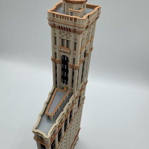 May include: A detailed model of a tall building with a unique, triangular shape. The building features a beige facade with brown trim, multiple windows, and a flat roof. The top of the building has a decorative tower.