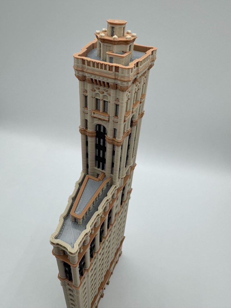 May include: A detailed model of a tall building with a unique, triangular shape. The building features a beige facade with brown trim, multiple windows, and a flat roof. The top of the building has a decorative tower.