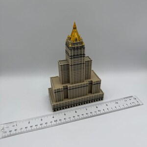 May include: A miniature model of a skyscraper with a gold-colored spire. The building is beige with dark window details. A clear ruler with inch measurements is placed next to the building.