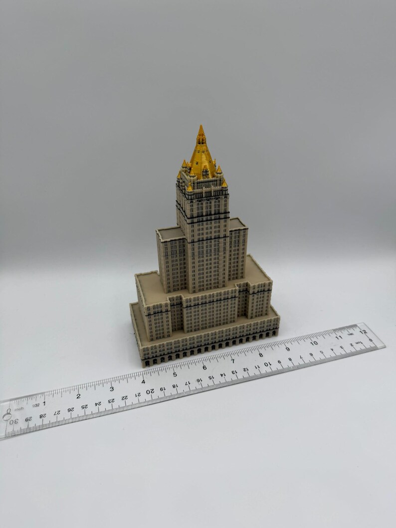 May include: A miniature model of a skyscraper with a gold-colored spire. The building is beige with dark window details. A clear ruler with inch measurements is placed next to the building.