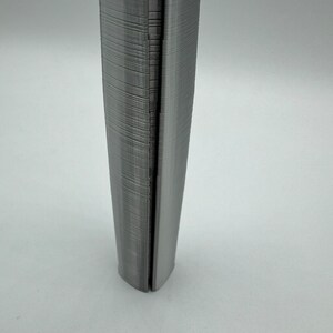 May include: A close-up of a gray, 3D printed object with a textured surface. The object is shaped like a cylinder and has a slight curve at the bottom.