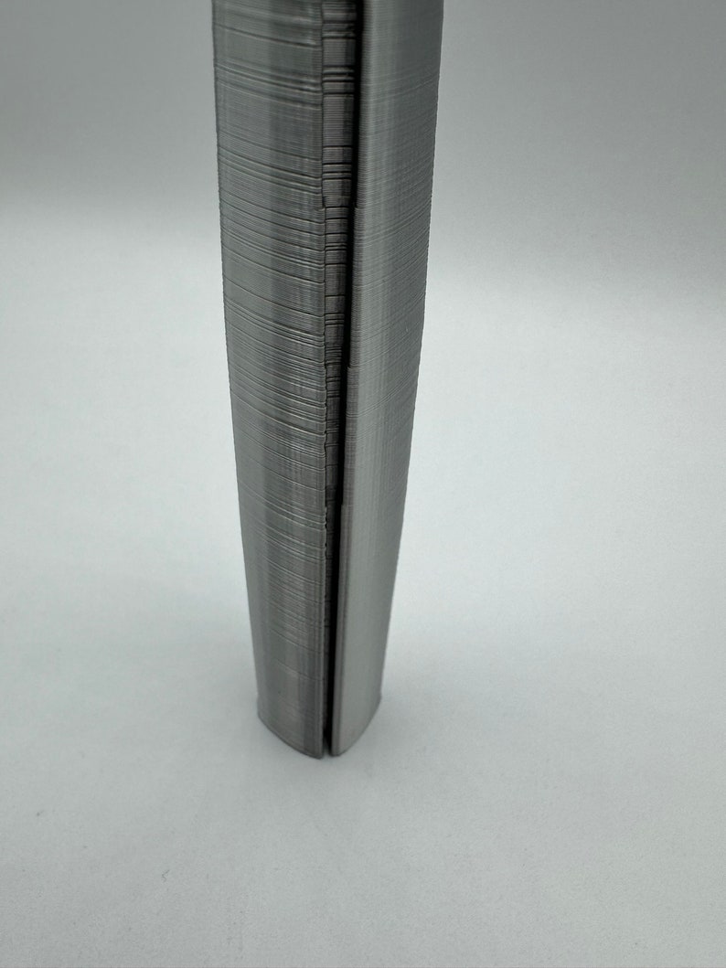 May include: A close-up of a gray, 3D printed object with a textured surface. The object is shaped like a cylinder and has a slight curve at the bottom.