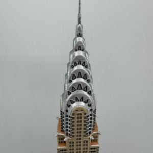 May include: A 3D-printed model of the Chrysler Building in New York City. The model features a silver spire and Art Deco design, with a tan base and silver upper levels. The building's windows are visible, and the model is set against a plain background.