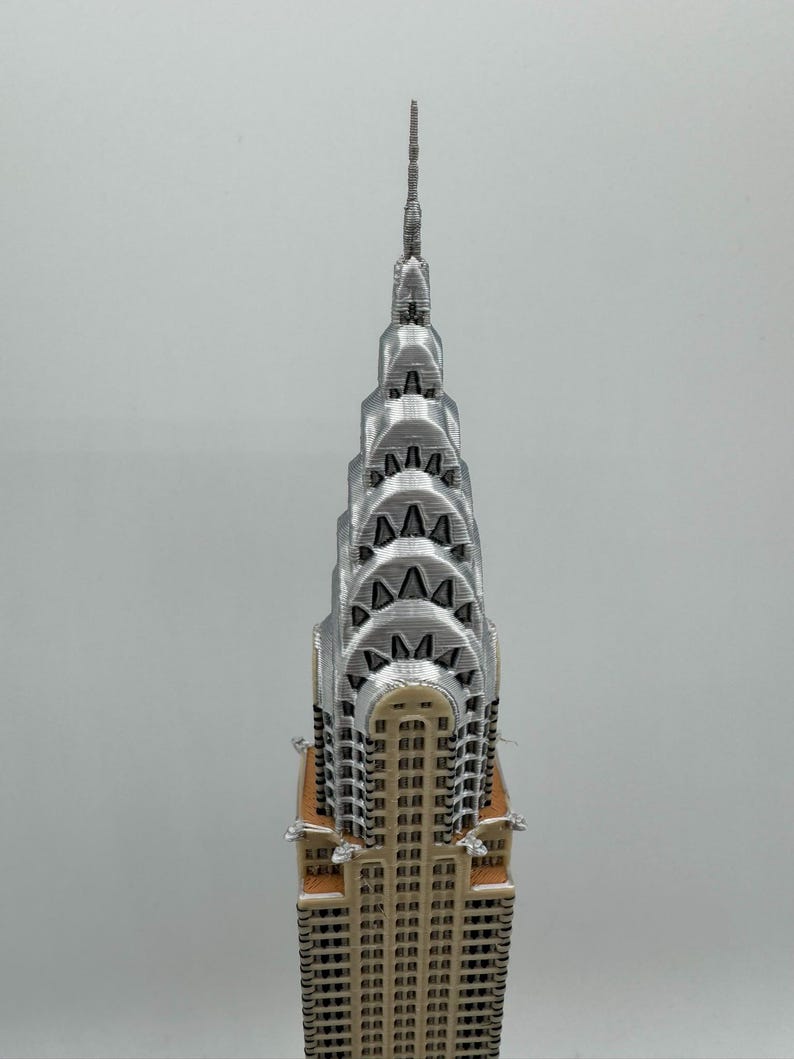 May include: A 3D-printed model of the Chrysler Building in New York City. The model features a silver spire and Art Deco design, with a tan base and silver upper levels. The building's windows are visible, and the model is set against a plain background.