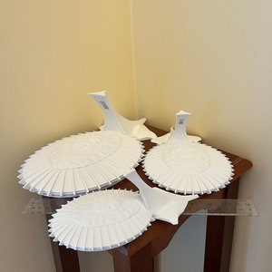 May include: Three white ceramic cake stands with a scalloped edge and a raised center. The stands are stacked on top of each other on a wooden table.