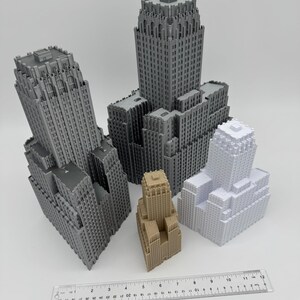 May include: A collection of miniature skyscraper models in various shades of gray, beige, and white. A ruler is placed at the bottom, showing measurements in inches. The buildings have detailed architectural features.