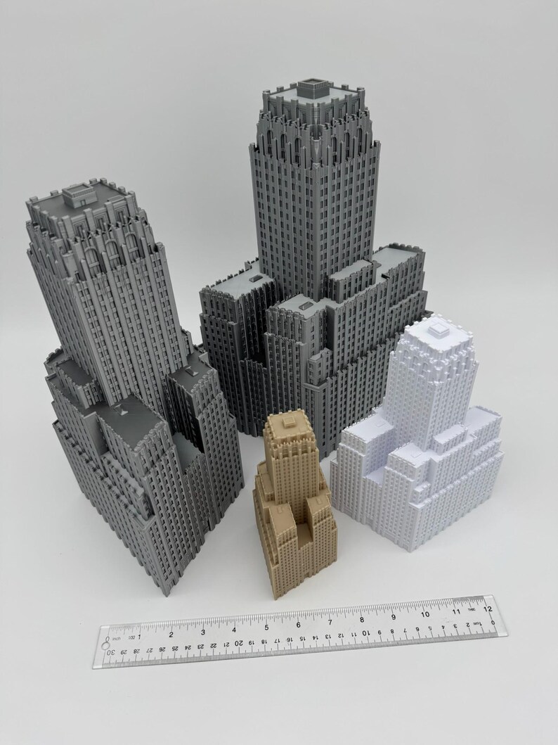 May include: A collection of miniature skyscraper models in various shades of gray, beige, and white. A ruler is placed at the bottom, showing measurements in inches. The buildings have detailed architectural features.