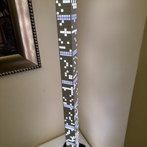 May include: A tall, rectangular floor lamp designed to resemble a skyscraper. The lamp features a gray exterior with illuminated white and blue square patterns, creating a pixelated effect. It stands on a black square base and has a white cord.