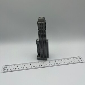 May include: A gray miniature model of a tall skyscraper building, approximately 6 inches tall, sitting on a white surface next to a ruler.