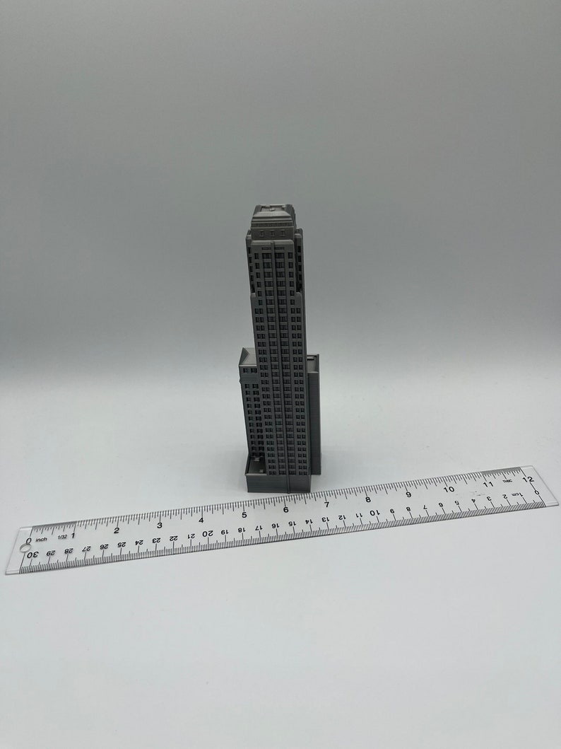 May include: A gray miniature model of a tall skyscraper building, approximately 6 inches tall, sitting on a white surface next to a ruler.