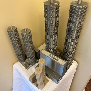 May include: A set of five 3D printed models of skyscrapers. The models are made of a light gray plastic and are arranged on a white tablecloth. The models are all different sizes and shapes, and some have intricate details.