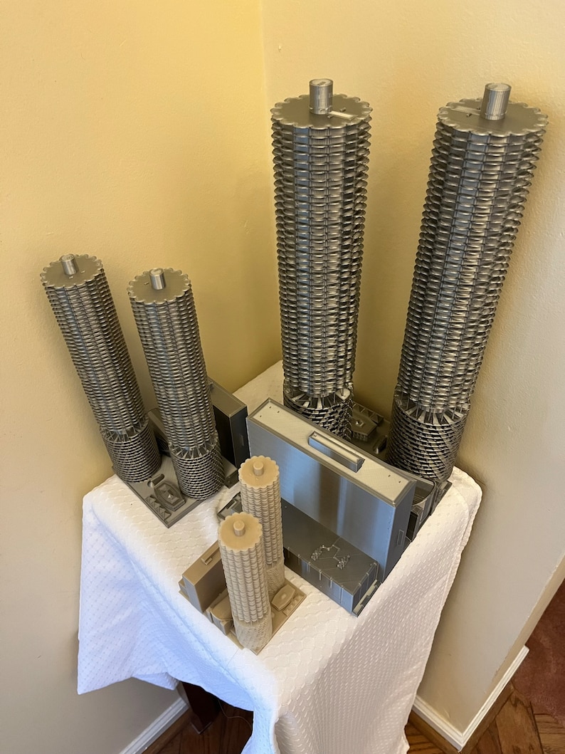 May include: A set of five 3D printed models of skyscrapers. The models are made of a light gray plastic and are arranged on a white tablecloth. The models are all different sizes and shapes, and some have intricate details.