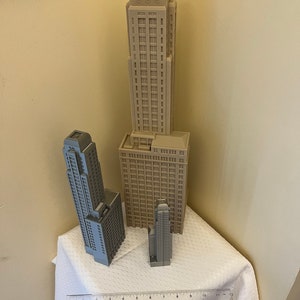 May include: Three miniature building models, two are gray and one is beige, on a white tablecloth with a ruler in the foreground. The tallest building is beige and has a flat roof with a decorative design. The two gray buildings are taller than the ruler, which is 12 inches long.