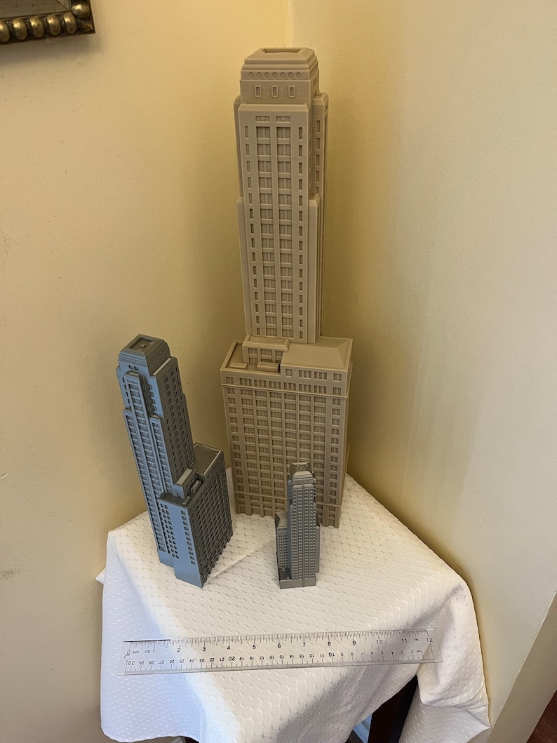 May include: Three miniature building models, two are gray and one is beige, on a white tablecloth with a ruler in the foreground. The tallest building is beige and has a flat roof with a decorative design. The two gray buildings are taller than the ruler, which is 12 inches long.