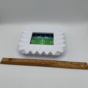 May include: A white paper stadium model with a green field and blue seating. The stadium is a geometric design with a detailed field and goal lines. A wooden ruler is placed next to the stadium.