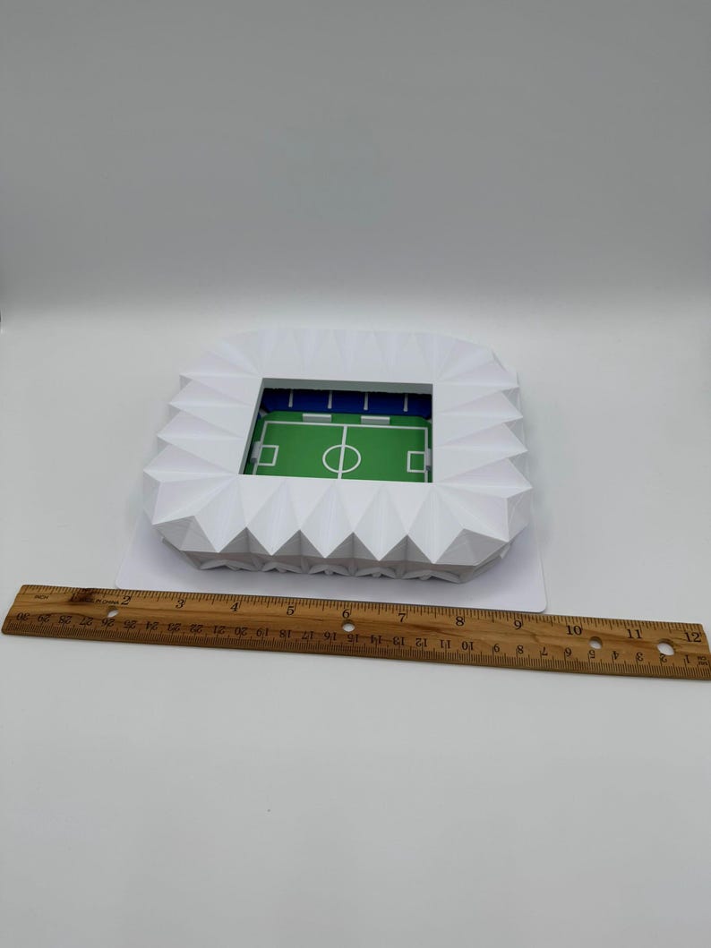 May include: A white paper stadium model with a green field and blue seating. The stadium is a geometric design with a detailed field and goal lines. A wooden ruler is placed next to the stadium.