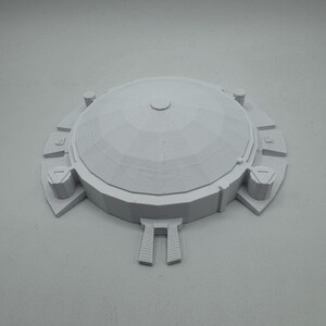 May include: A light gray, 3D-printed model of a domed structure. The model features a central dome with concentric rings, surrounded by a circular base with rectangular and triangular architectural elements. The overall design is symmetrical.