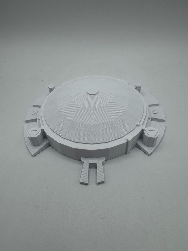 May include: A light gray, 3D-printed model of a domed structure. The model features a central dome with concentric rings, surrounded by a circular base with rectangular and triangular architectural elements. The overall design is symmetrical.