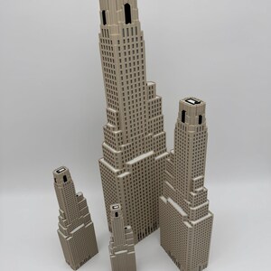 May include: A set of four beige skyscraper models with tiered architectural designs and black window details. A wooden ruler is placed at the base, showing the tallest model reaches approximately 30.5 cm in height, providing a clear scale reference.