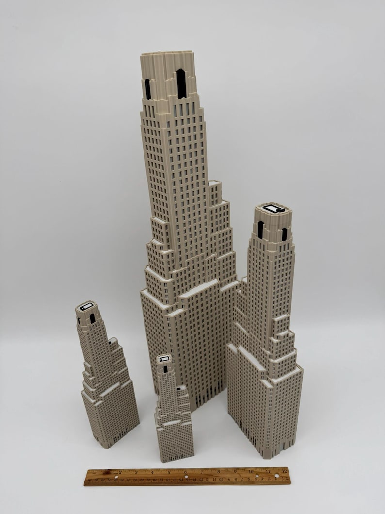 May include: A set of four beige skyscraper models with tiered architectural designs and black window details. A wooden ruler is placed at the base, showing the tallest model reaches approximately 30.5 cm in height, providing a clear scale reference.