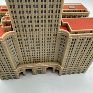 May include: A detailed model of a beige-colored skyscraper with numerous windows and a red roof. The building has a classic architectural design, with arched entryways and a tall, rectangular structure. The model is likely a collectible or decorative item.