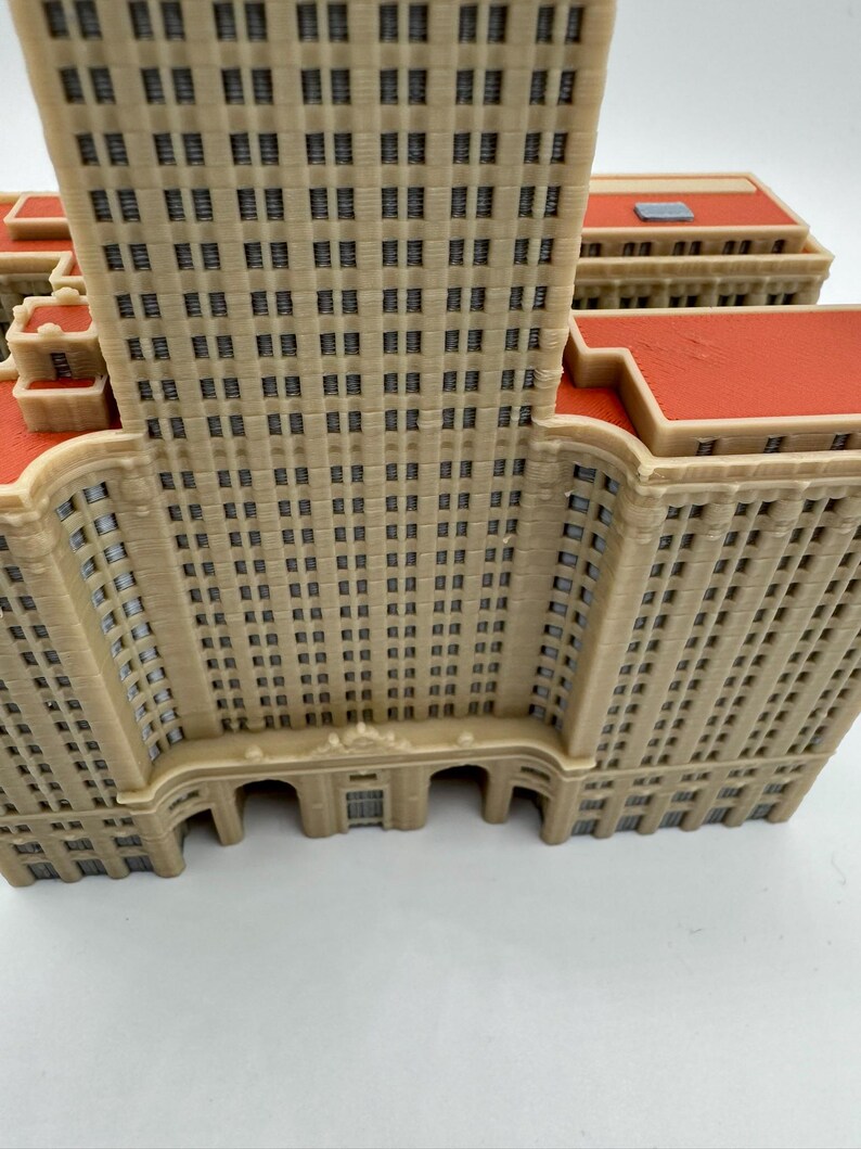 May include: A detailed model of a beige-colored skyscraper with numerous windows and a red roof. The building has a classic architectural design, with arched entryways and a tall, rectangular structure. The model is likely a collectible or decorative item.