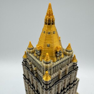 May include: A detailed 3D-printed model of a building with a gold-colored spire and accents. The building has a beige facade with black trim and multiple levels. The top of the building is gold, with several gold-colored spires.