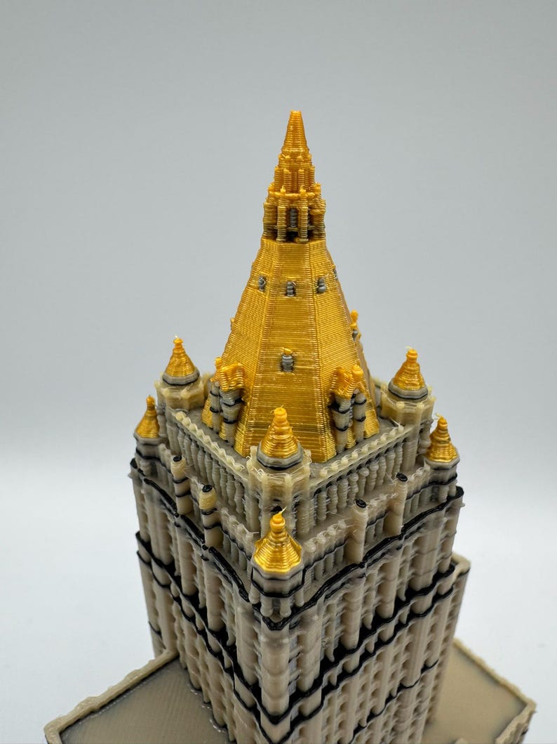 May include: A detailed 3D-printed model of a building with a gold-colored spire and accents. The building has a beige facade with black trim and multiple levels. The top of the building is gold, with several gold-colored spires.