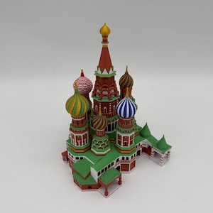 May include: A miniature model of Saint Basil's Cathedral featuring a central red tower with a gold spire, surrounded by multiple onion domes in various colors including yellow, green, red, white, and blue stripes, set on a green base.