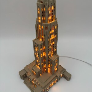 May include: A beige, 3D-printed architectural model of a tall skyscraper with a stepped design. The structure features numerous rectangular windows that emit a warm, glowing orange light from within, creating a realistic illuminated effect.